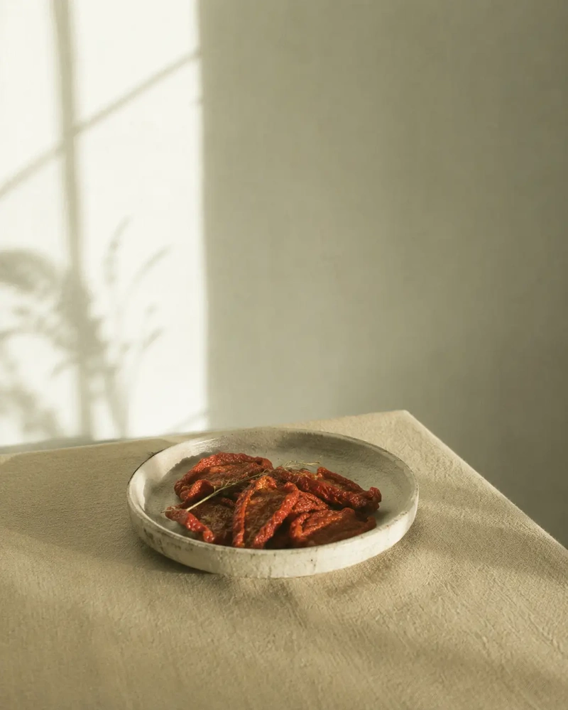 Umami foods Minimalist still life of sun-dried tomatoes arranged in a shallow ceramic plate, photographed from a slightly different angle on a pale textured surface with soft natural light and a warm neutral background.