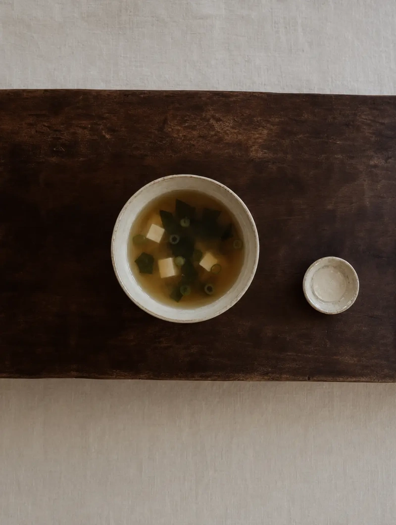 Umami Foods Overhead minimalist composition of a ceramic bowl filled with miso broth on a dark wooden board, accompanied by a small empty dish, styled in soft neutral tones with a calm editorial mood.