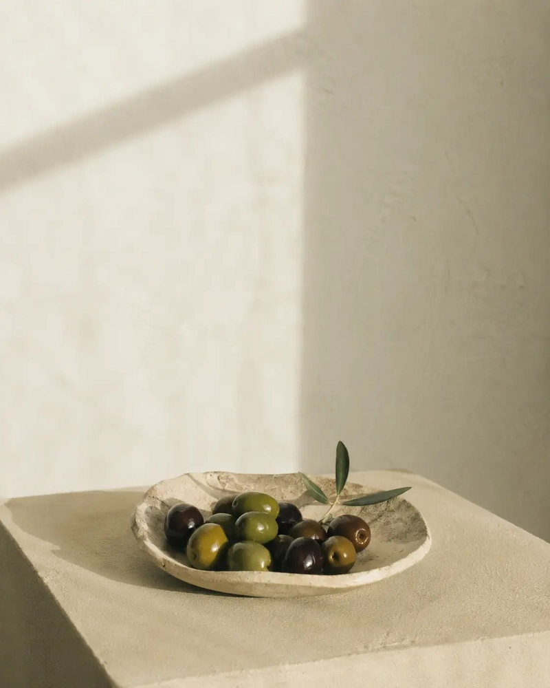 Umami Foods Minimalist still life of mixed green and black olives in a shallow ceramic plate, styled on a pale textured pedestal with soft sunlight and warm shadows against a neutral wall.