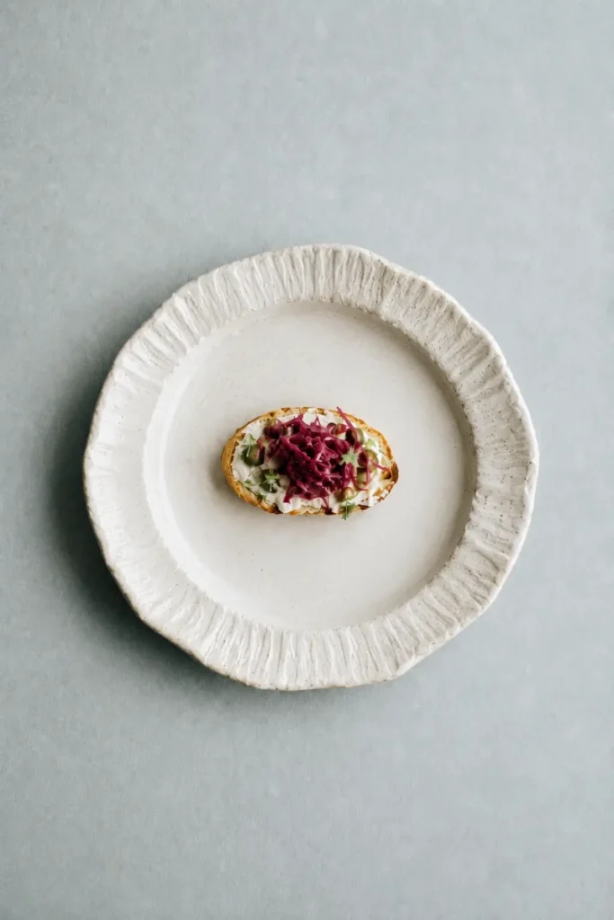 Fermented beets served on a small tartine with creamy spread and herbs on a textured white ceramic plate.
