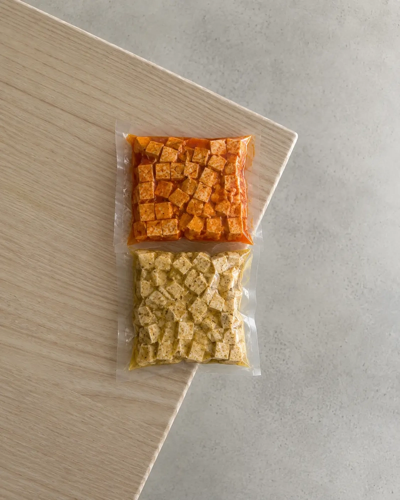 Marinated tofu cubes sealed in two vacuum bags with orange and yellow marinades on a pale wooden board and neutral grey background.
