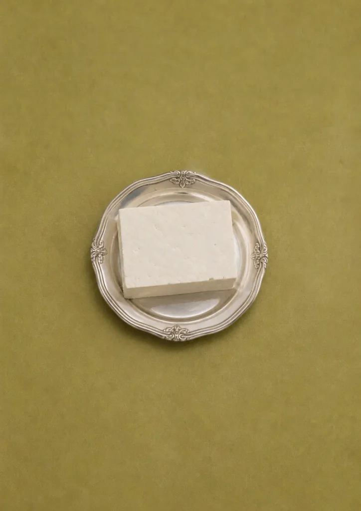 How to press tofu without a tofu press: block of tofu on a vintage silver plate against an olive green background in a minimal overhead composition.