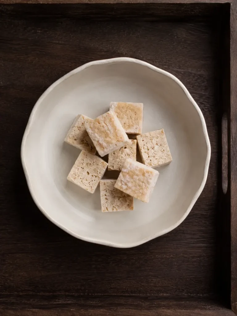 Frozen tofu cubes on a ceramic bowl, served on a dark wooden tray in a minimal overhead food composition.