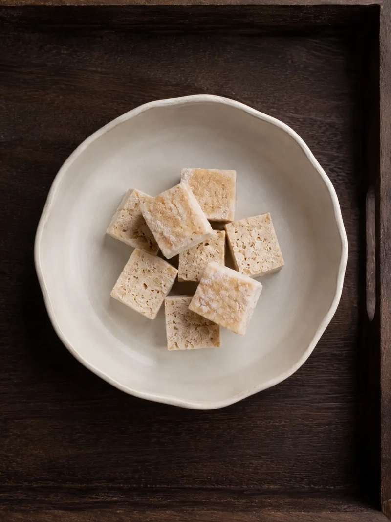 Frozen tofu cubes on a ceramic bowl, served on a dark wooden tray in a minimal overhead food composition.