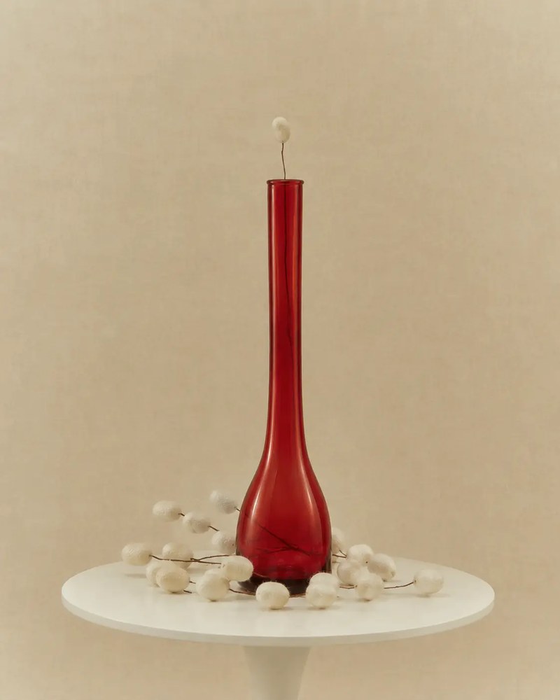 Is silk vegan article image with a red glass vase surrounded by white silkworm cocoons on a minimalist table against a warm beige backdrop.