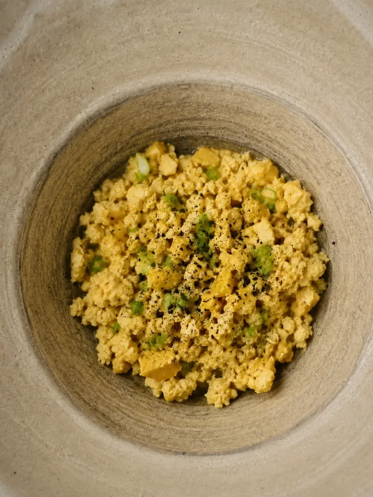 Creamy vegan scrambled eggs served in a rustic beige ceramic bowl, finished with fine kala namak black salt and delicate green scallion slices.