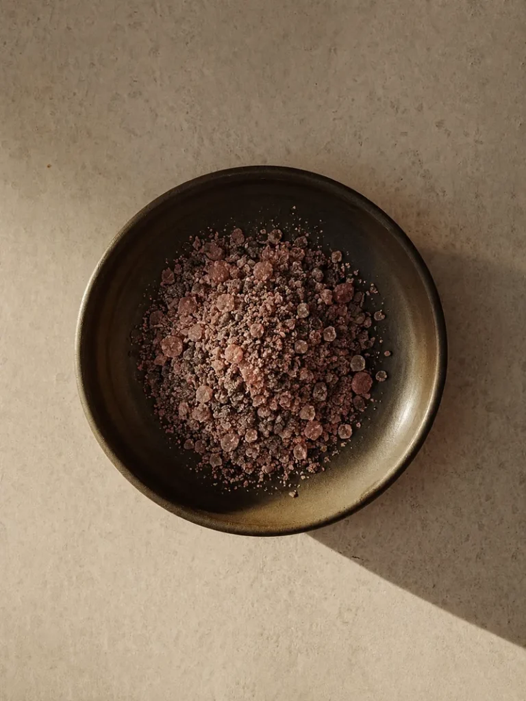 Pile of coarse kala namak black salt crystals served in a dark ceramic bowl on a warm stone surface, photographed from above with soft natural shadows.