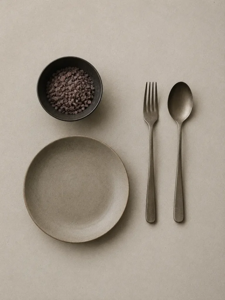Kala namak salt in a black bowl, styled beside an empty ceramic plate, fork, and spoon on a neutral textured surface.