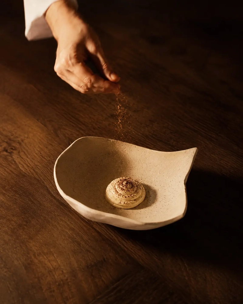 A hand sprinkling kala namak black salt over vegan mayonnaise served in an elegant handmade ceramic dish on a warm wooden table.