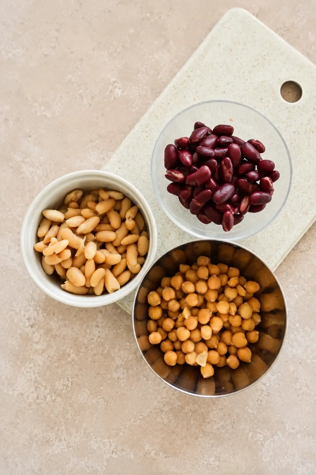 Three bowls of beans for a vegan bean salad, with chickpeas, cannellini beans, and kidney beans arranged on a pale cutting board.