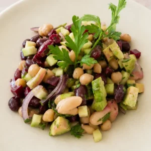 Close-up of a fresh vegan bean salad with mixed beans, cucumber, avocado, beetroot, red onion, and parsley on a pale ceramic plate.