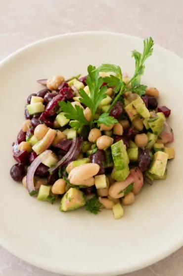 Close-up of a fresh vegan bean salad with mixed beans, cucumber, avocado, beetroot, red onion, and parsley on a pale ceramic plate.