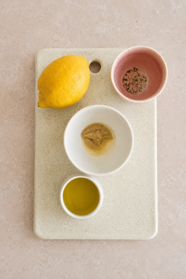 Vegan bean salad dressing ingredients with lemon, olive oil, mustard, salt, and black pepper on a pale cutting board.