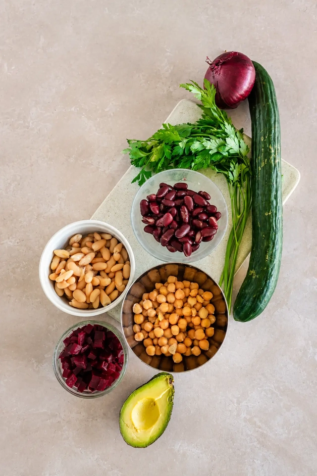 Vegan bean salad ingredients with chickpeas, cannellini beans, kidney beans, beetroot, avocado, cucumber, parsley, and red onion on a neutral kitchen surface.
