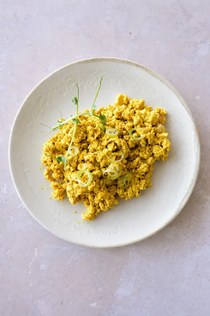Vegan scrambled eggs made with tofu, served in a ceramic bowl with green onion rings and microgreens on a neutral stone background.