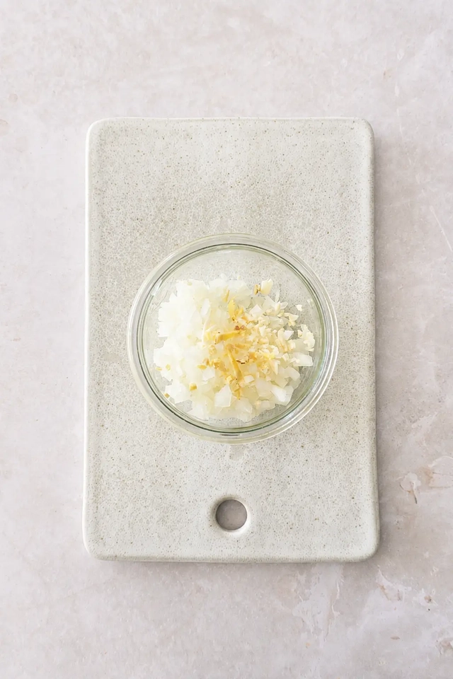 Chopped onion and garlic in a small glass bowl placed on a speckled off-white cutting board, photographed from above on a pale stone surface.