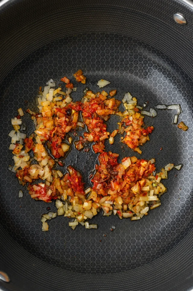 Vegan korma chopped onion and tomato paste sautéing in oil inside a large nonstick pan, photographed from above.