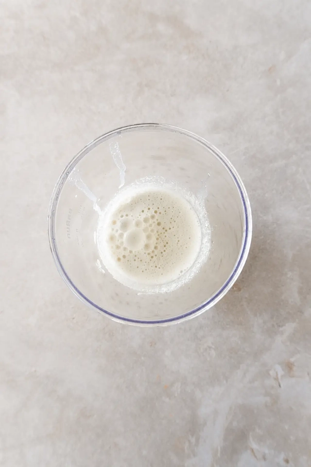 vegan Korma Creamy blended cashew mixture with small bubbles inside a clear blender cup, photographed from above on a neutral stone surface.