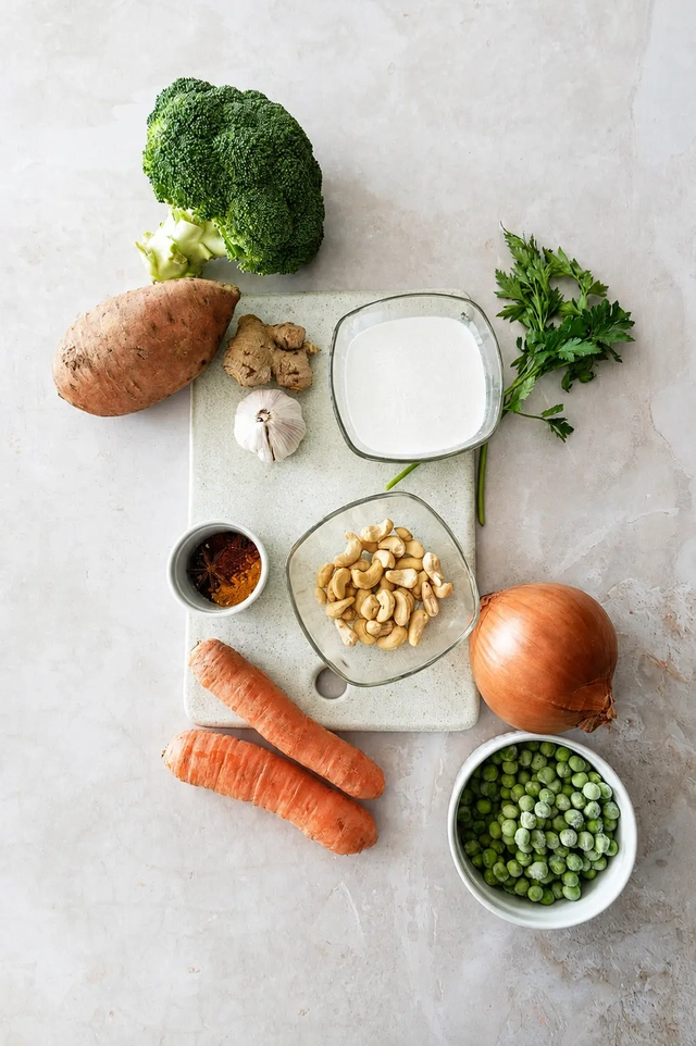 Vegan korma recipe ingredients arranged on a neutral surface with broccoli, sweet potato, carrots, peas, onion, garlic, ginger, cashews, coconut milk, parsley, and spices