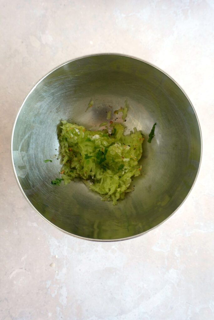 Vegan tzatziki sauce recipe step showing squeezed cucumber mixed with fresh mint, salt, and black pepper in a stainless steel bowl.