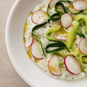 Vegan tzatziki sauce recipe served in a white bowl with cucumber ribbons, radish slices, olive oil, black pepper, and fresh herbs.