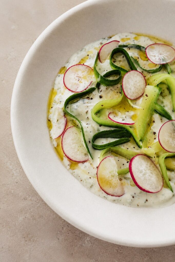 Vegan tzatziki sauce recipe served in a white bowl with cucumber ribbons, radish slices, olive oil, black pepper, and fresh herbs.