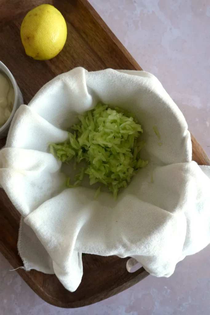 Vegan tzatziki sauce recipe step showing grated cucumber placed in a clean kitchen towel before being squeezed to remove excess water.