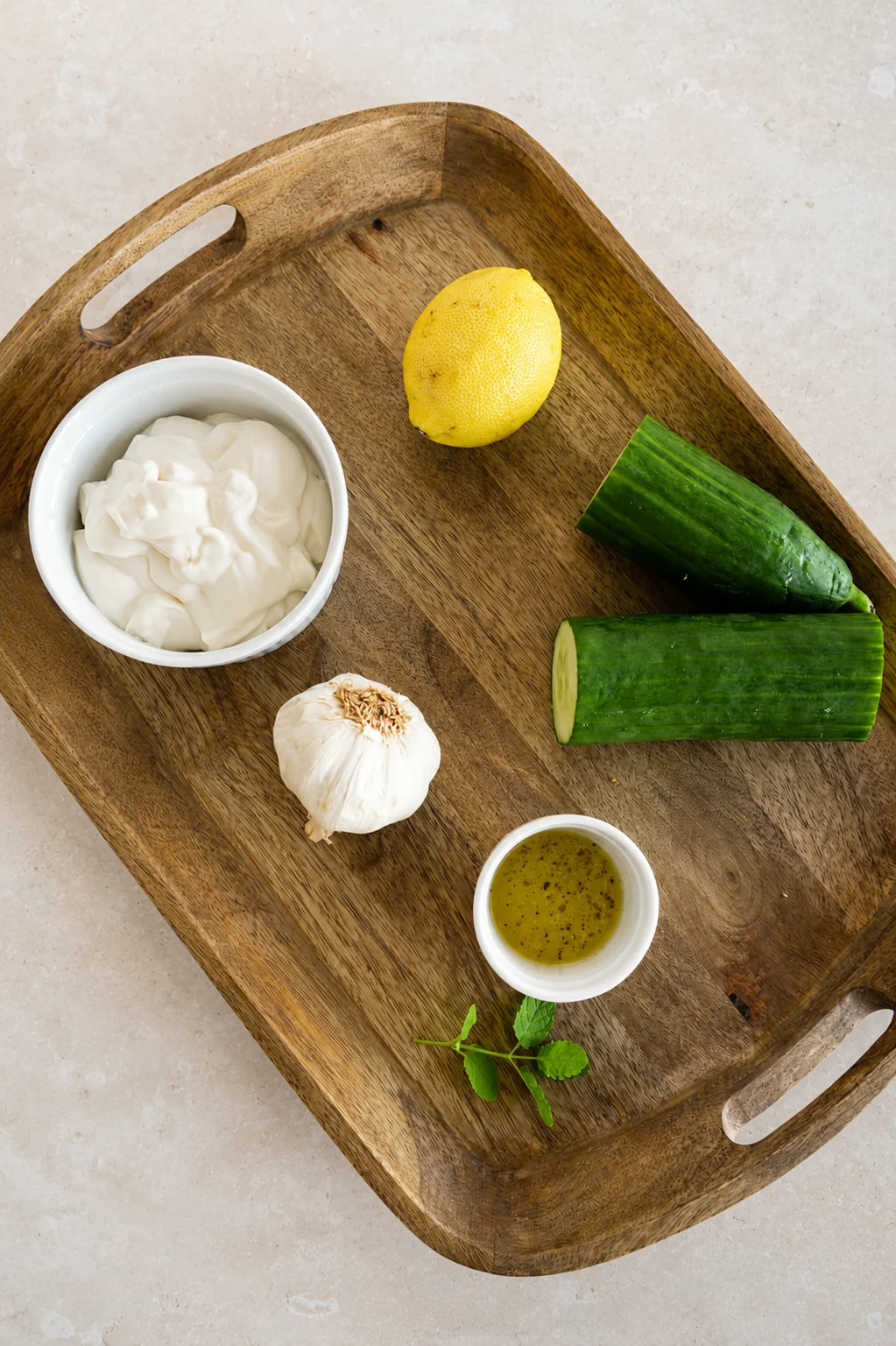 Vegan tzatziki sauce recipe ingredients arranged on a wooden tray, including soy yogurt, cucumber, lemon, garlic, olive oil, and fresh mint.