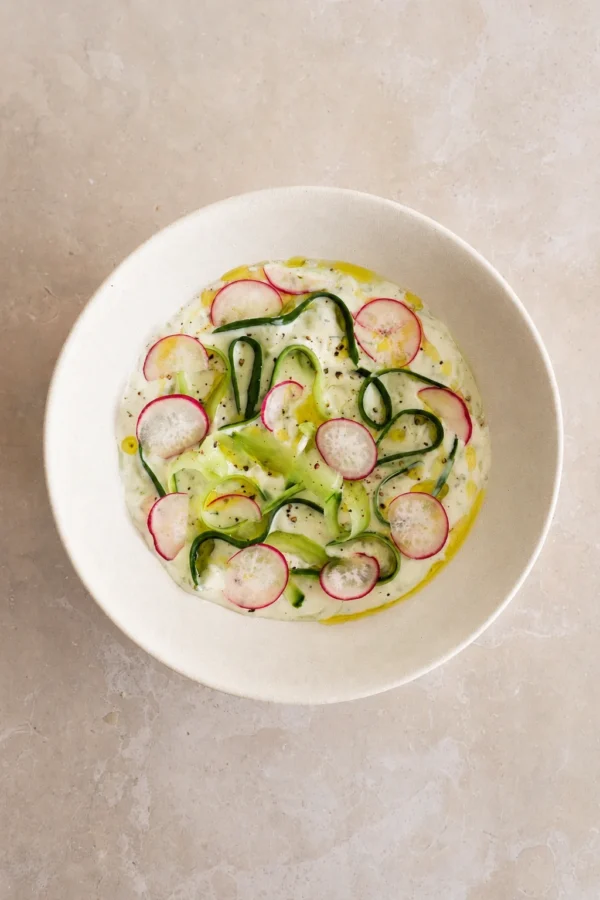 Vegan tzatziki sauce recipe served in a white bowl with cucumber ribbons, radish slices, olive oil, black pepper, and fresh mint.
