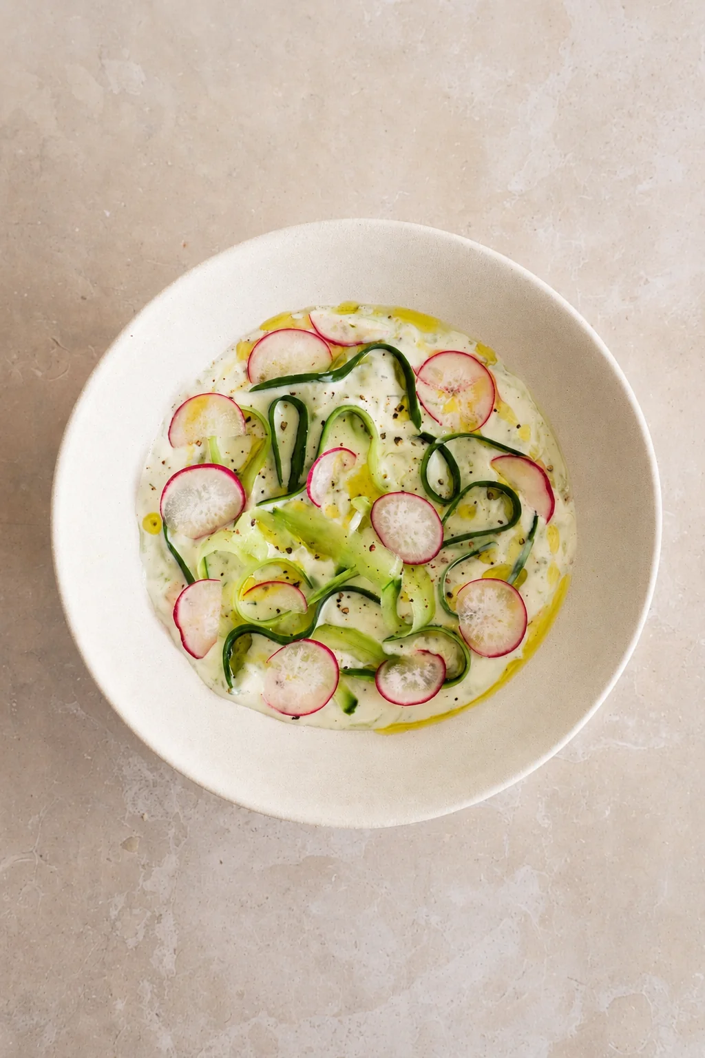 Vegan tzatziki sauce recipe served in a white bowl with cucumber ribbons, radish slices, olive oil, black pepper, and fresh mint.