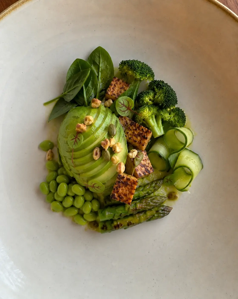 Tempeh bowl with grilled tempeh cubes, fanned avocado, broccoli, edamame, asparagus, cucumber ribbons, spinach, and seeds, plated in a refined gourmet style on a wide ceramic bowl.