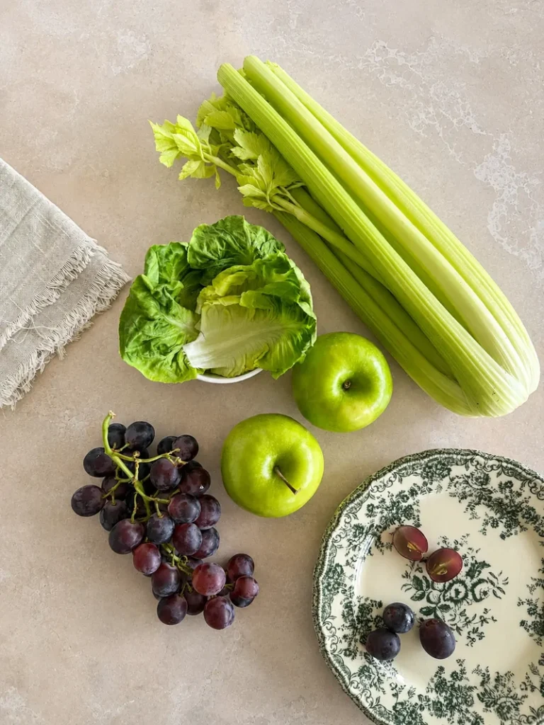 Vegan Waldorf Salad ingredients arranged in a bright overhead flat lay, featuring celery, green apples, red grapes, crisp lettuce leaves, and a decorative plate on a light stone surface.