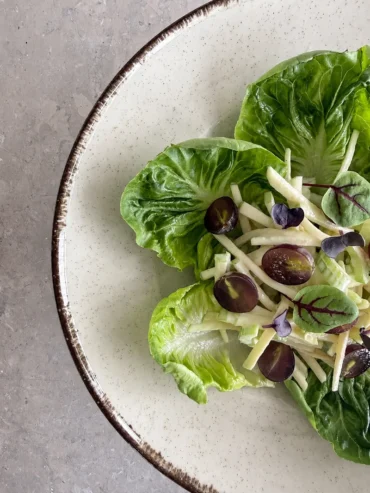 Vegan Waldorf Salad served over crisp lettuce leaves with creamy julienned apple and celery, halved red grapes, and delicate microgreens on a ceramic white plate.