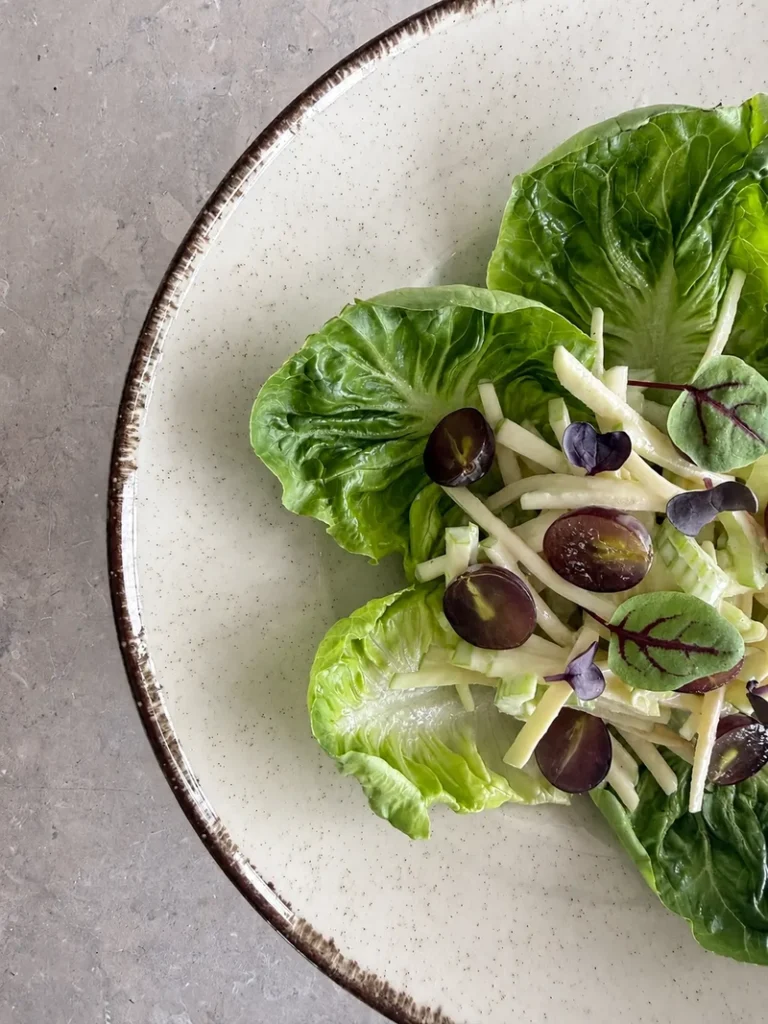 Vegan Waldorf Salad shown in a close-up editorial composition with crisp lettuce leaves, creamy julienned apple and celery, halved red grapes, and delicate microgreens on a ceramic plate.
