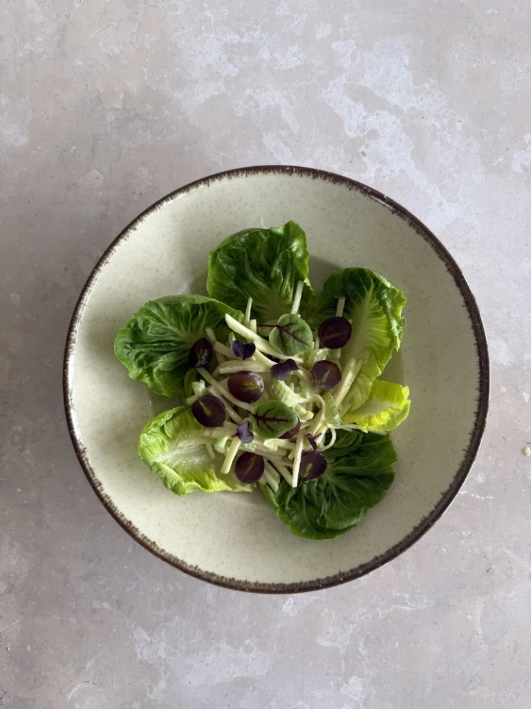 Vegan Waldorf Salad served over crisp lettuce leaves with creamy julienned apple and celery, halved red grapes, and delicate microgreens on a ceramic plate.