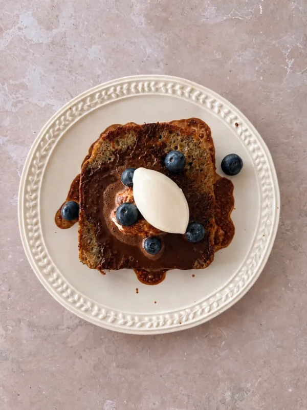 Vegan French toast served on a white plate, topped with creamy vegan yogurt quenelle, fresh blueberries, and a rich chocolate drizzle.