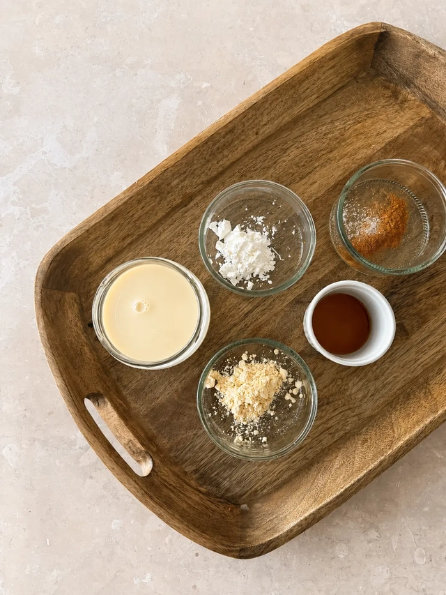 Ingredients for vegan French toast arranged on a wooden tray, including plant-based milk, chickpea flour, cornstarch, vanilla extract, cinnamon, and salt in small bowls, photographed from above.