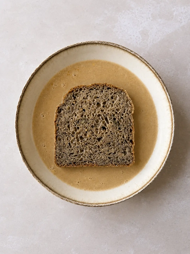 Thick slice of homemade buckwheat bread placed in vegan French toast batter before soaking, photographed from above in a ceramic bowl.