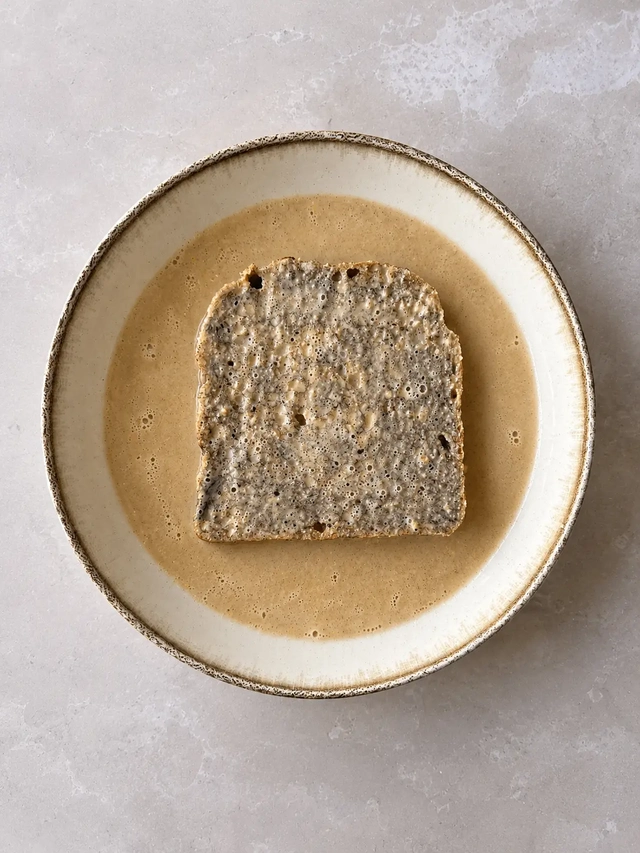 Slice of gluten-free buckwheat bread soaking in vegan French toast batter in a rustic ceramic bowl, photographed from above.