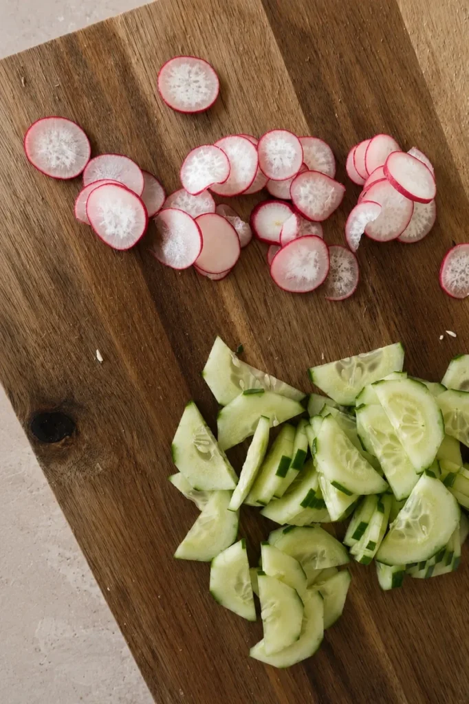 Sliced radishes and cucumber on a wooden cutting board before assembling the salad