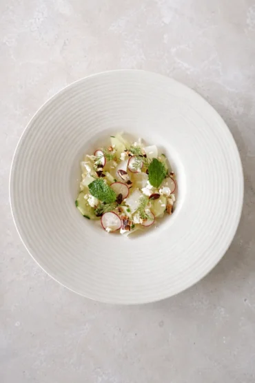 Honeydew cucumber salad with lime-marinated melon, vegan feta, radishes, pecans, mint, and fresh dill served on a white ceramic plate.