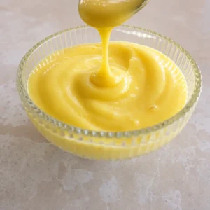 Close-up of thick, glossy vegan lemon curd being poured from a spoon into a small ribbed glass bowl on a light stone surface.