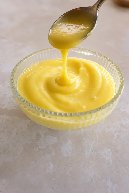 Close-up of thick, glossy vegan lemon curd being poured from a spoon into a small ribbed glass bowl on a light stone surface.