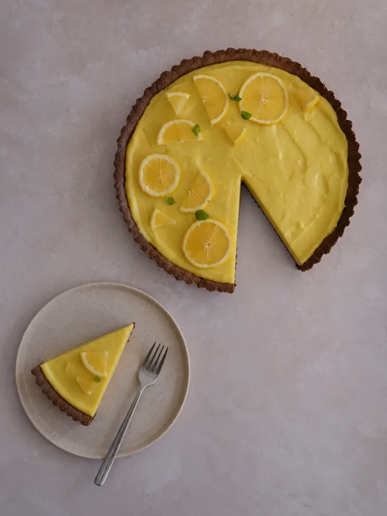 Overhead view of a vegan lemon tart with a gluten-free crust, topped with lemon slices and mint leaves, with one slice removed and served on a small plate with a fork on a neutral stone background.