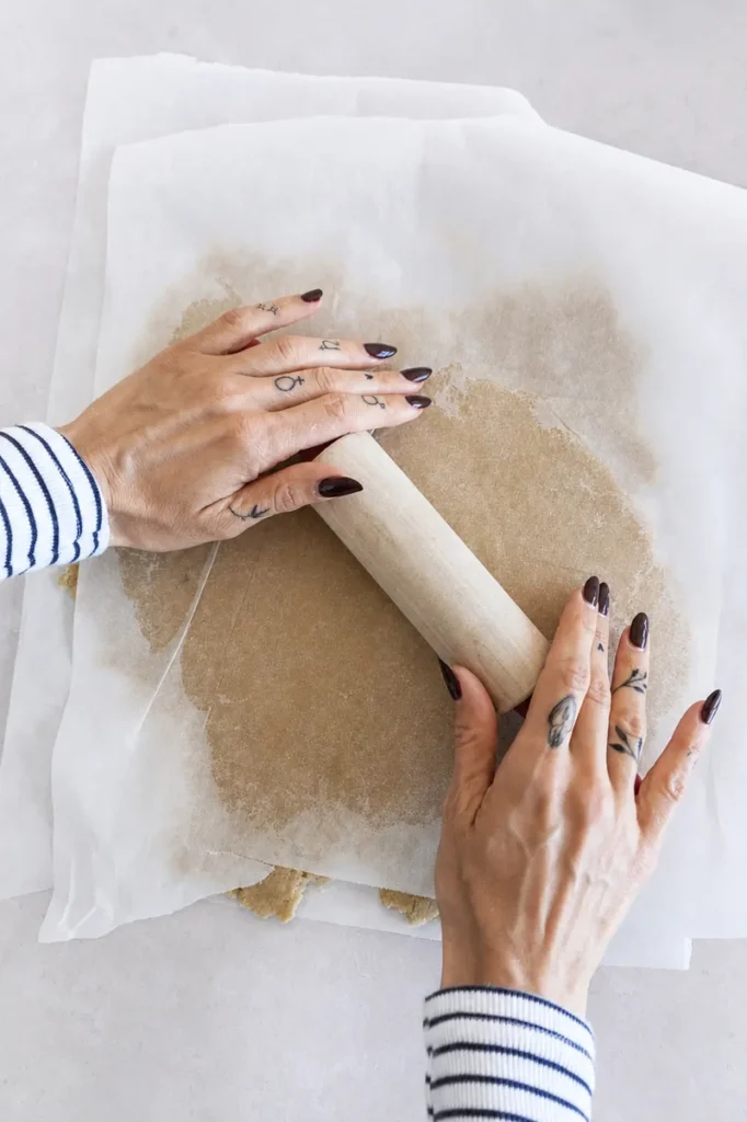 Hands rolling the gluten-free crust for a vegan lemon curd tart between sheets of parchment paper with a wooden rolling pin.