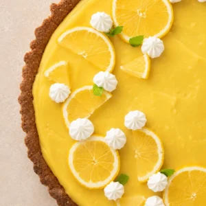 Close-up of a vegan lemon curd tart with a smooth lemon filling, lemon slices, mint leaves and neat white cream dollops piped with a pastry bag.