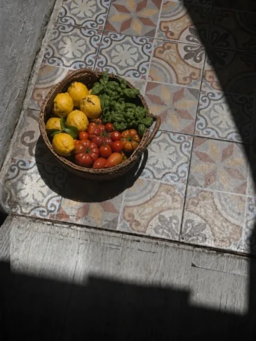 Vegan Mediterranean diet ingredients in a rustic basket with lemons, mixed tomatoes, and fresh basil on patterned Mediterranean floor tiles in natural sunlight.