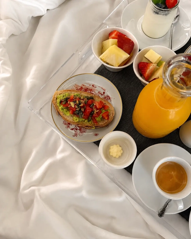 What I eat in a day as a vegan breakfast with avocado toast, fresh fruit, plant-based yogurt, orange juice, and coffee served on a tray in bed.
