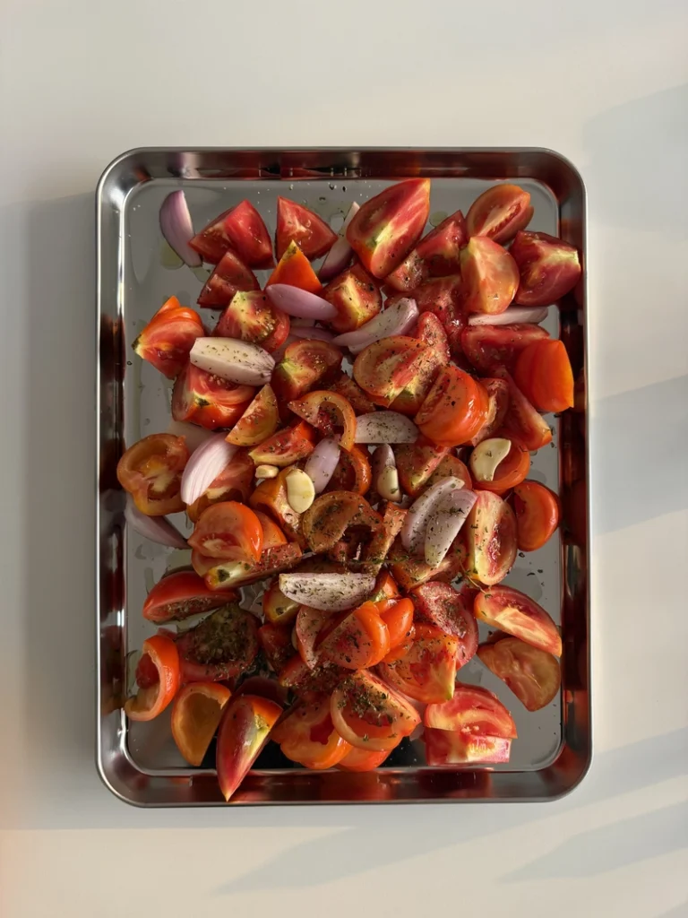 Fresh tomatoes, red onion and garlic seasoned with olive oil, herbs, salt and pepper on a baking tray before roasting for 3-ingredient tomato soup.