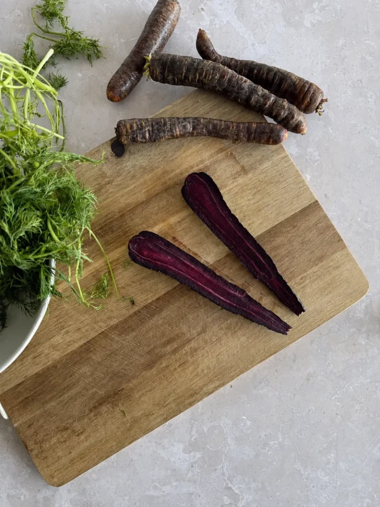 Heirloom purple carrots cut in half on a wooden board, with fresh carrot tops and contrasting purple and orange flesh.